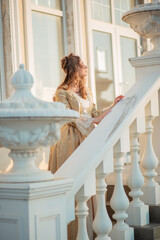 A beautiful young woman in a historic eighteenth century gold dress stands on the stairs of the...
