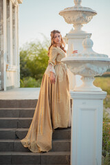 Fototapeta premium A beautiful young woman in a historic eighteenth century gold dress stands on the stairs of the mansion. Princess in the palace.