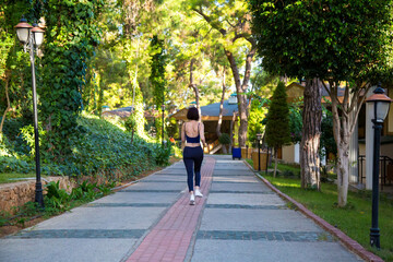 female athlete runs along the sidewalk in hotel along green palm trees firs and pines.