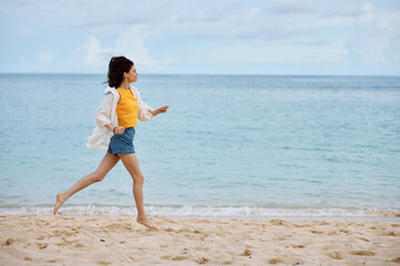 Fototapeta na wymiar Sports woman runs along the beach in summer clothes on the sand in a yellow tank top and denim shorts white shirt flying hair ocean view, beach vacation and travel