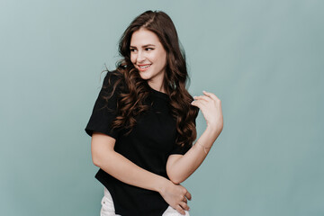 Attractive shy brunette girl toothy smiling looking aside twirls her curly long hair. Hispanic model posing at studio over turquoise backdrop. Playful young woman dancing indoors. Beauty and fashion.