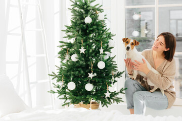 Smiling woman embraces pet with love, has happy mood, pose together near beautiful decorated...