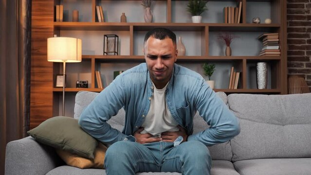 African American man sitting on sofa feeling stomach pain at home, gastritis symptom, peptic ulcer, pancreatitis. Male guy suffering from digestive problems on couch, squeezing belly abdominal pain.