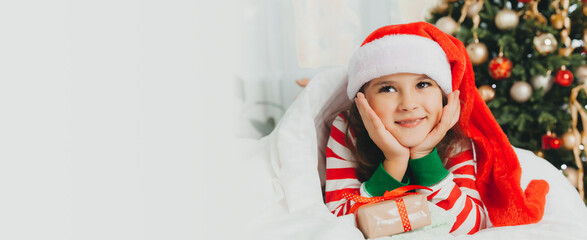 little girl in a New Year's red hat with a Christmas gift. She lies on the bed, hugging a box against the background of a Christmas tree.