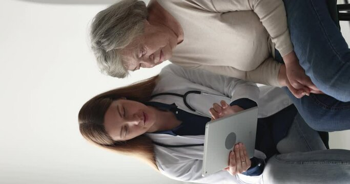 Woman general practitioner in white coat show lab test health result on digital tablet to elderly 80s female patient at meeting in clinic, medical worker use modern tech, healthcare app, vertical view
