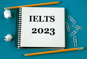 IELTS 2023 - word in a white notebook on a blue background with pencils, paper clips