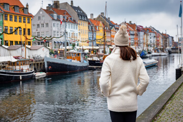 A tourist woman in winter clothing enjoys the view to the beautiful Nyhavn area in Copenhagen, Denmark, during winter time - obrazy, fototapety, plakaty