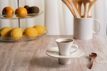 Fototapeta na wymiar Cup of coffee on a kitchen table with a fruit bowl and a jug with wooden utensils, in the background a white curtain.