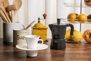 Cup of coffee on a kitchen table with a fruit bowl, a jug with wooden utensils, a coffee grinder and an Italian coffee maker, in the background a white curtain.