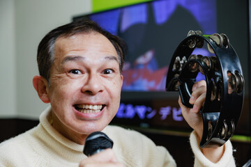 Middle-aged Asian man in white sweater with tambourine and microphone enjoying karaoke.