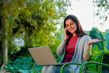 Young indian woman using laptop and talking on smartphone at park