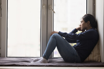 Unhappy lonely young Indian woman sitting on windowsill, looking out of window, thinking over bad news, loss, feeling sad, frustrated, suffering from apathy, depression. Side view