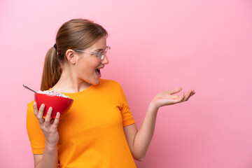 Young caucasian woman holding a bowl of cereals isolated on pink background with surprise facial...
