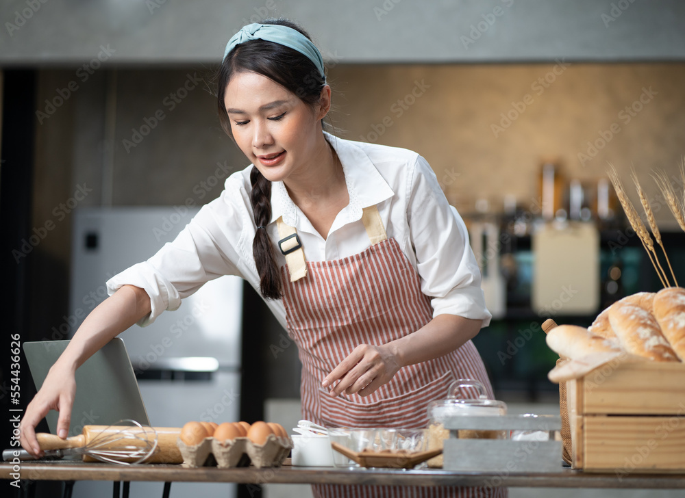 Wall mural happy asian female baker wears apron standing at table in kitchen preparing homemade bakery ingredie