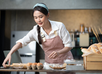 Happy Asian female baker wears apron standing at table in kitchen preparing homemade bakery ingredients. Smiling young beautiful woman baking bread and cooking at home as her small cookery business. - obrazy, fototapety, plakaty