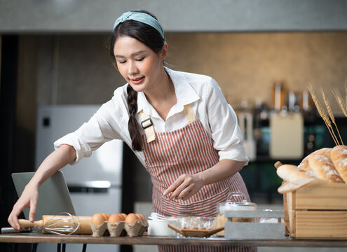 Happy Asian female baker wears apron standing at table in kitchen preparing homemade bakery ingredients. Smiling young beautiful woman baking bread and cooking at home as her small cookery business.