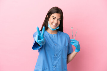 Dentist woman holding tools isolated on pink background smiling and showing victory sign