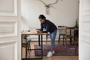 Happy millennial adult Indian girl working on business project in home office, standing at...