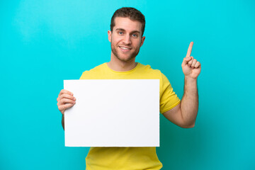 Young caucasian man isolated on blue background holding an empty placard and pointing up