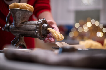 Nahaufnahme von Hände einer Frau, die mit einen alten Drehwolf, Spritzgebäck Plätzchen zu Weihnachten backt. Im Hintergrund das Bokeh von Weihnachtsbeleuchtung