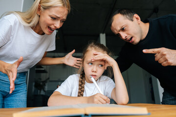 Portrait of tired little girl doing homework sitting at desk at home on background of angry parents...
