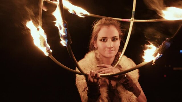 Attractive girl with plait, fur collar on neck, holding burning sphere formed with metal rods in front of her, smiles. Young actress demonstrates trick in fire show with round ignited props at night.