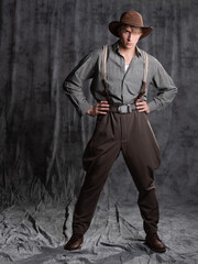 A young man in retro style, an adventure character. A guy in a hat and a gray shirt, breeches with suspenders. - 554450447