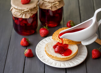 Canning Strawberries. Adding homemade strawberry syrup on sliced white bread bun with portion of whole strawberry berries on it. Selective focus. Glass jars with canned strawberries on background.