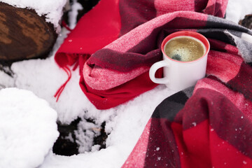 A mug of hot coffee wrapped in a warm checkered scarf on snow-covered bars.
