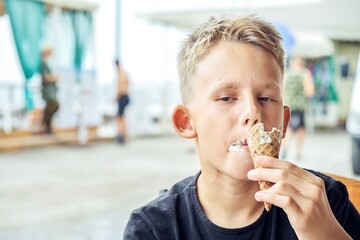 Junior schoolboy enjoys eating ice cream and spending summer holidays at sea resort. Blond-haired boy eats ice cream with satisfied expression - 554455201
