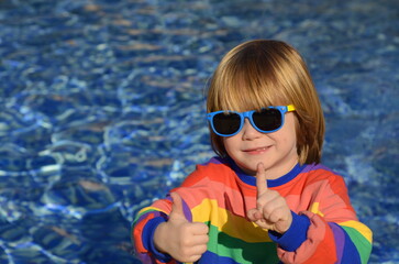 Positive little boy in sunglasses and a multi-colored jacket on the background of the water. Space for text, sun glare on the surface of the pool. Concept: resort, fountain, traveling with children