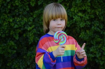 Funny kid with caramel on a stick. Cute positive boy 3 years old in a colorful t-shirt. Against the background of green grass. Space for text. Concept: kindergarten, harmful candies, holidays, good mo