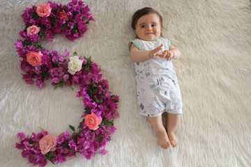 Cute baby lie on back on white fluffy blanket. Shape of the number five with bougainvillea and rose...