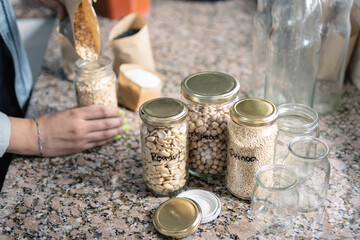 Table in the kitchen with bulk food in recycled jars and woman behind taking groceries from paper bags. Zero waste at home concept