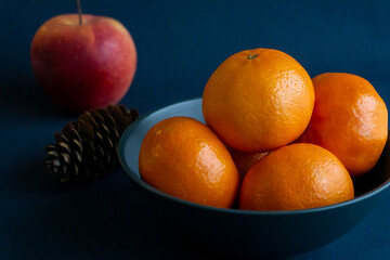 Fresh tangerines in a bowl, cone and an apple in the background. Christmas fruits. Dark background, closeup image.