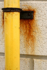 A bright yellow drain pipe with rust dripping off of it, set against the left side of a white wall. Image has copy space on the top, bottom and right side.