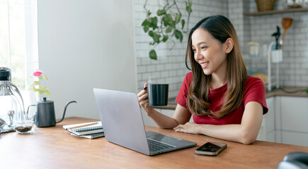 Beautiful young woman working on laptop computer while sitting at the kitchen room, drinking coffee