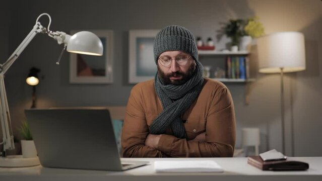 Low air temperature at home. Freezing bearded man in hat and scarf is trying to keep warm. Cold in apartment, in house, indoors