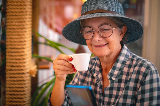 Handsome senior woman with hat and eyeglasses relaxes in cafeteria holding a coffee cup and looking at her mobile phone. Elderly lady enjoying free time and retirement