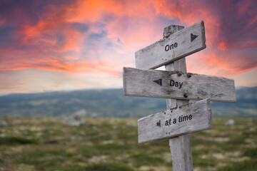 one day at a time text quote engraved on wooden signpost crossroad outdoors in nature. Dramatic pink skies in the background.