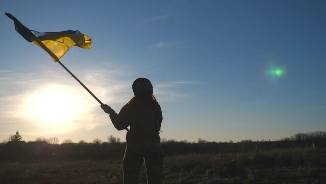Woman in military uniform waving flag of Ukraine against blue sky with sun. Female ukrainian army soldier lifted up flag. Victory against Russian aggression. Invasion resistance concept. Slow motion