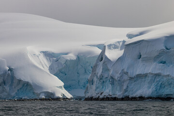 Closeup view of the edge of Antarctic Peninsula, revealing small inlet. Rocks covered with deep layers of snow, blue and white ice. Clouds in background; calm sea in foreground.

