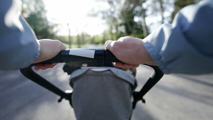 Hands holding stroller POV of mother walking outside in nature with baby. Parent pushing toddler carriage at park