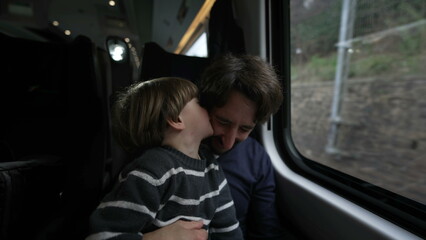 Son kissing father on cheek while traveling by train. Parent and child passengers inside moving high speed train. Kid showing love and affection