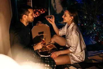 Fotobehang Valentines day home date ideas Save Money Budget Tips. Young couple in love celebrating Valentines day at home. Romantic couple in pajamas eating pizza near fireplace © irissca