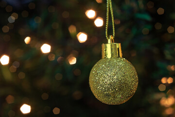 Uma bola dourada, pendurada no galho com detalhes da árvore de Natal e luzes ao fundo.