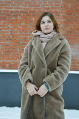 Naklejka premium A young pretty girl in a beige fur coat without a hat stands near a brick wall