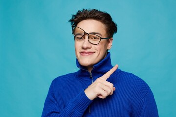  man stands on a light blue background in a blue zip-up sweater with black-rimmed glasses turned...
