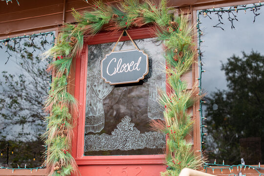 A glass decorative red wooden door of a shop with white curtains painted on the glass window. The store building has Christmas lights hanging around the windows. A closed sign hangs in the storefront.