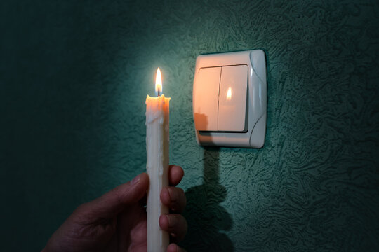 A man's hand holding a candle in total darkness near a light switch. 
 Power outage, blackout, concept of non-payment of power bill, energy crisis or blackout. Problems with power supply. 